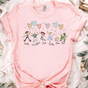 May include: A light pink t-shirt featuring a graphic of cartoon characters holding Mickey Mouse-shaped balloons. The characters include Woody, Buzz Lightyear, Jessie, Bo Peep, Hamm, and Rex. The names of the characters are printed below them.