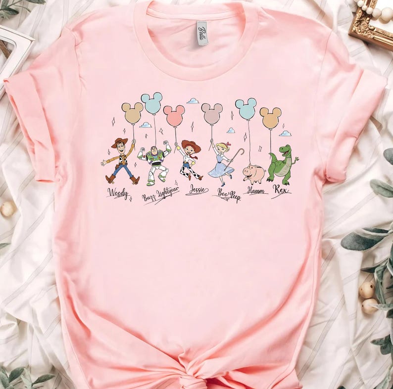 May include: A light pink t-shirt featuring a graphic of cartoon characters holding Mickey Mouse-shaped balloons. The characters include Woody, Buzz Lightyear, Jessie, Bo Peep, Hamm, and Rex. The names of the characters are printed below them.