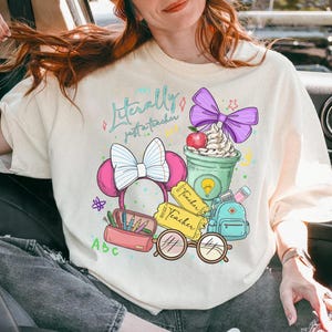 May include: Cream-colored t-shirt with a colorful graphic that reads "Literally just a teacher." The design includes a pink Minnie Mouse bow, a green coffee cup with whipped cream, a red apple, and school-themed elements.