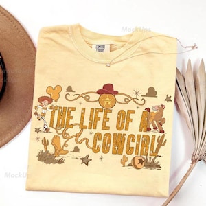May include: A light yellow t-shirt with the phrase "THE LIFE OF A COWGIRL" in gold lettering. The shirt features cartoon graphics of a cowgirl, a horse, and other western-themed elements. A brown hat is visible in the upper left corner.