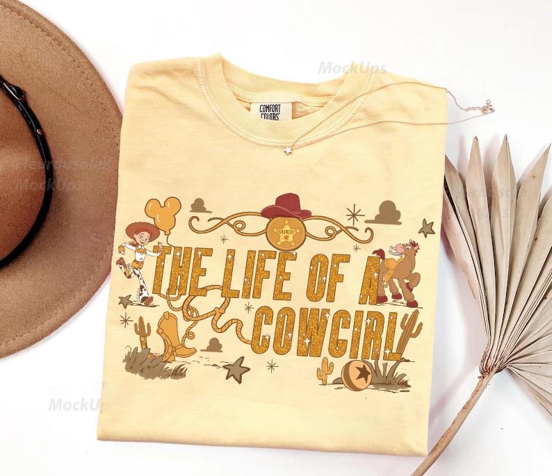 May include: A light yellow t-shirt with the phrase "THE LIFE OF A COWGIRL" in gold lettering. The shirt features cartoon graphics of a cowgirl, a horse, and other western-themed elements. A brown hat is visible in the upper left corner.