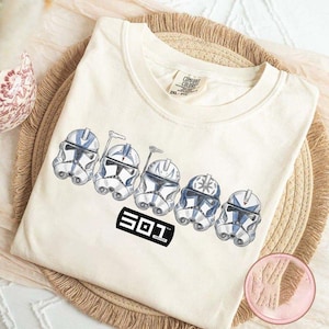 May include: Cream-colored t-shirt featuring a graphic of five white and blue clone trooper helmets. The text "501" is printed in a black rectangle below the helmets. The shirt is folded on a woven mat.
