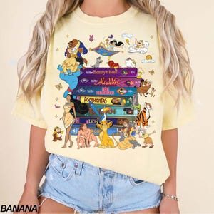 Puede incluir: Camiseta amarilla clara con una impresi&oacute;n de t&iacute;tulos y personajes de pel&iacute;culas de animaci&oacute;n, incluyendo La Bella y la Bestia, Aladdin y El Rey Le&oacute;n. La camiseta tiene cuello redondo y mangas cortas. La palabra "BANANA" est&aacute; impresa en la parte inferior izquierda.