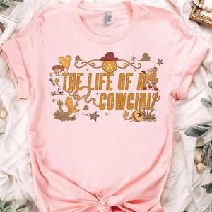 May include: A light pink t-shirt featuring the text "THE LIFE OF A COWGIRL" in gold lettering. The design includes cartoon illustrations of a cowgirl, cowboy boots, a horse, and other western-themed elements.