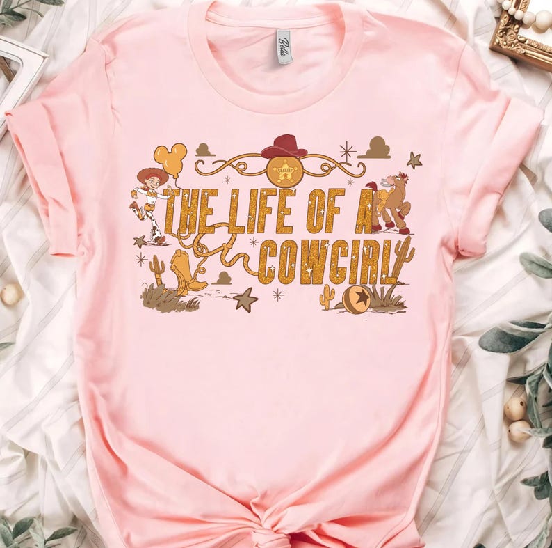 May include: A light pink t-shirt featuring the text "THE LIFE OF A COWGIRL" in gold lettering. The design includes cartoon illustrations of a cowgirl, cowboy boots, a horse, and other western-themed elements.