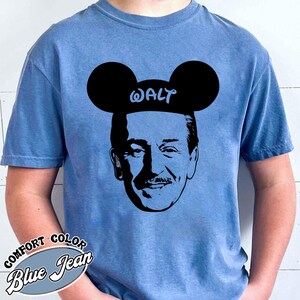 Puede incluir: Camiseta azul vaquero Comfort Colors con una silueta negra de Walt Disney con orejas de Mickey Mouse con el nombre "WALT" en las orejas. La camiseta tiene las palabras "COMFORT COLOR Blue Jean" impresas en la parte inferior izquierda.