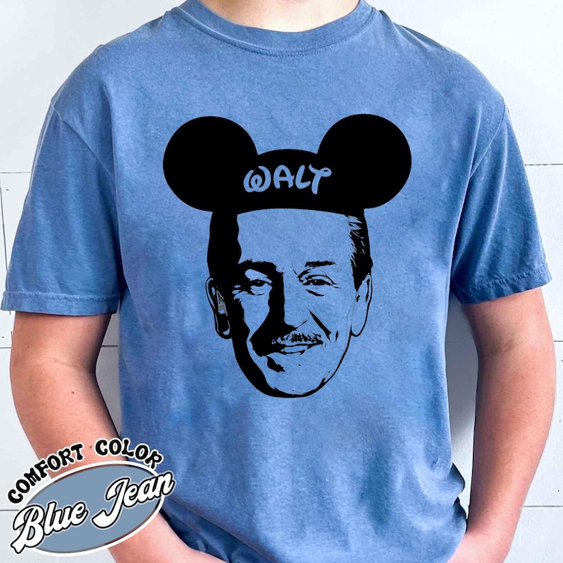 Puede incluir: Camiseta azul vaquero Comfort Colors con una silueta negra de Walt Disney con orejas de Mickey Mouse con el nombre "WALT" en las orejas. La camiseta tiene las palabras "COMFORT COLOR Blue Jean" impresas en la parte inferior izquierda.