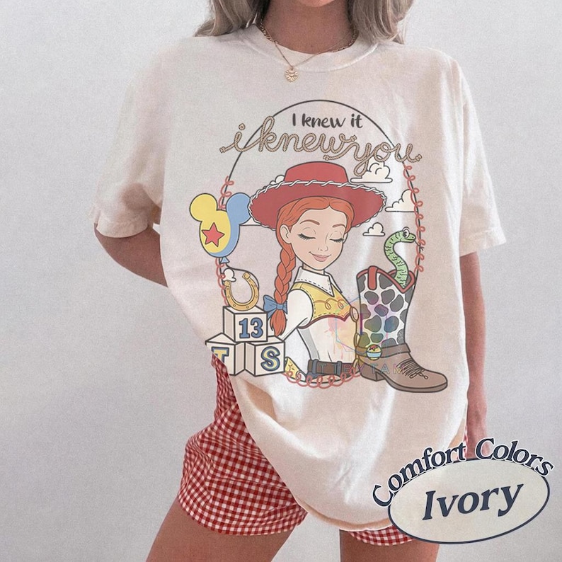 May include: An ivory t-shirt featuring a graphic of a cowgirl character with red hair, a red hat, and a yellow vest. The design includes the text I knew it i knew you, a balloon, a horseshoe, alphabet blocks with 13, and a patterned cowboy boot.