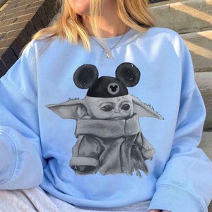 May include: A light blue crewneck sweatshirt featuring a graphic print of a small, grey, large-eared creature wearing a black cap with round mouse ears. The cap displays a small, circular icon with a mouse ear silhouette in the center.