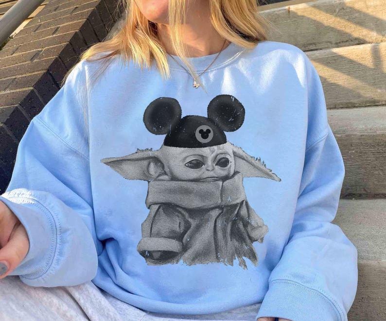 May include: A light blue crewneck sweatshirt featuring a graphic print of a small, grey, large-eared creature wearing a black cap with round mouse ears. The cap displays a small, circular icon with a mouse ear silhouette in the center.