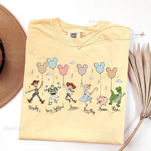 May include: A light yellow t-shirt featuring cartoon characters holding Mickey Mouse-shaped balloons. The characters include Woody, Buzz Lightyear, Jessie, Bo Peep, Hamm, and Rex. The shirt has the words "Comfort Colors" on the tag.
