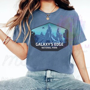 Puede incluir: Una camiseta azul con un dise&ntilde;o hexagonal que muestra un paisaje monta&ntilde;oso y el texto "GALAXY'S EDGE NATIONAL PARK". El dise&ntilde;o incluye una escena futurista con un veh&iacute;culo volador.