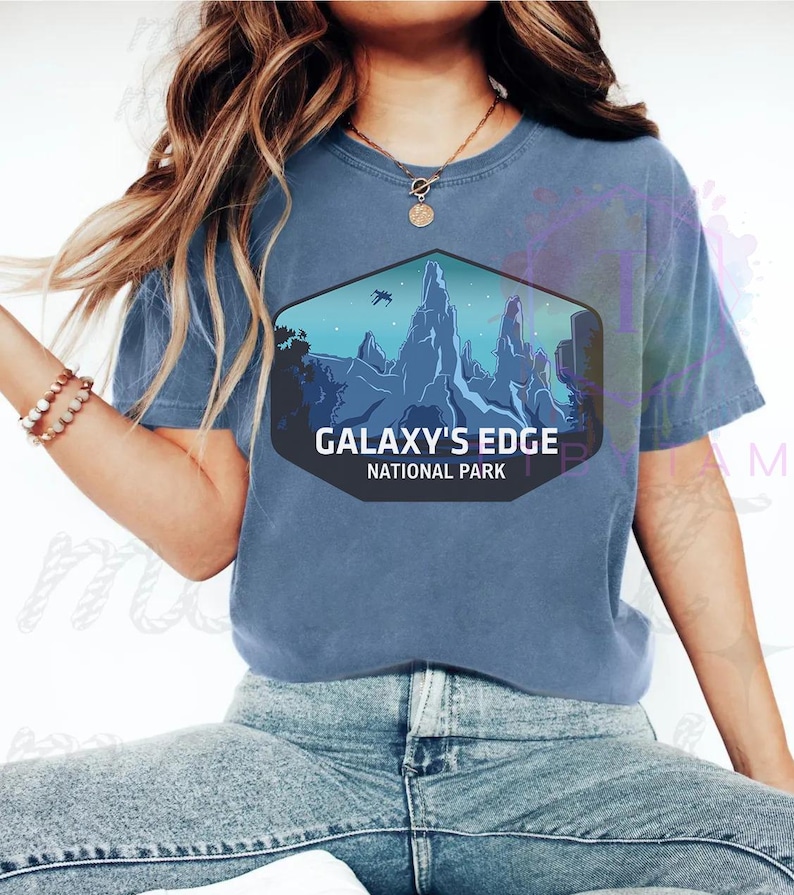 Puede incluir: Una camiseta azul con un dise&ntilde;o hexagonal que muestra un paisaje monta&ntilde;oso y el texto "GALAXY'S EDGE NATIONAL PARK". El dise&ntilde;o incluye una escena futurista con un veh&iacute;culo volador.
