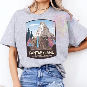 Puede incluir: Camiseta gris claro con un gr&aacute;fico de un paisaje monta&ntilde;oso, una cascada, &aacute;rboles y el texto "FANTASYLAND - NATIONAL PARK" en un marco marr&oacute;n. La camiseta est&aacute; hecha de un material suave.