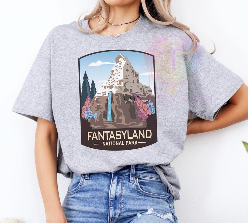 Puede incluir: Camiseta gris claro con un gr&aacute;fico de un paisaje monta&ntilde;oso, una cascada, &aacute;rboles y el texto "FANTASYLAND - NATIONAL PARK" en un marco marr&oacute;n. La camiseta est&aacute; hecha de un material suave.