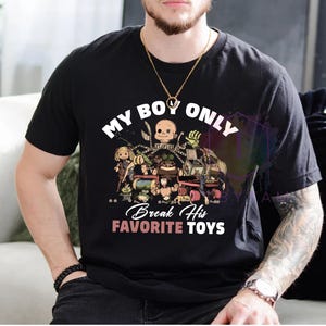 K&ouml;nnte beinhalten: Ein schwarzes Kurzarm-T-Shirt mit einem Grafikdruck verschiedener Spielzeugfiguren. Der Text lautet MY BOY ONLY Break His FAVORITE TOYS in wei&szlig;er und rosa Schrift. Das Design zeigt unter anderem einen Puppenkopf und eine Spieluhr mit Figur.