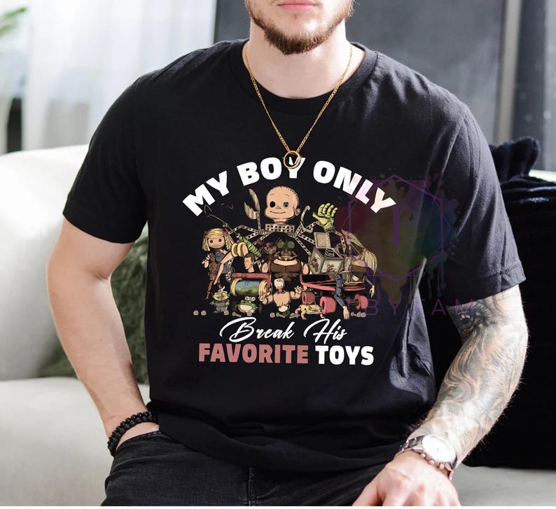K&ouml;nnte beinhalten: Ein schwarzes Kurzarm-T-Shirt mit einem Grafikdruck verschiedener Spielzeugfiguren. Der Text lautet MY BOY ONLY Break His FAVORITE TOYS in wei&szlig;er und rosa Schrift. Das Design zeigt unter anderem einen Puppenkopf und eine Spieluhr mit Figur.