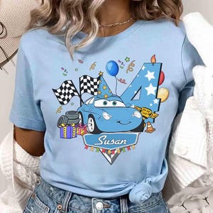 May include: Light blue t-shirt featuring a cartoon car with a birthday theme. The design includes a number 4, checkered flags, balloons, and the name "Susan". The car has a party hat and is surrounded by festive elements.