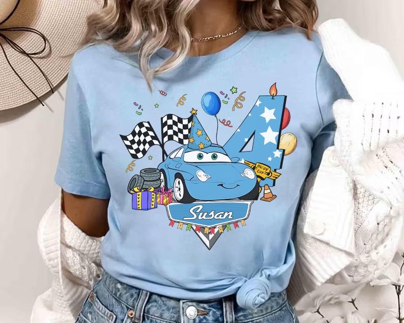 May include: Light blue t-shirt featuring a cartoon car with a birthday theme. The design includes a number 4, checkered flags, balloons, and the name "Susan". The car has a party hat and is surrounded by festive elements.
