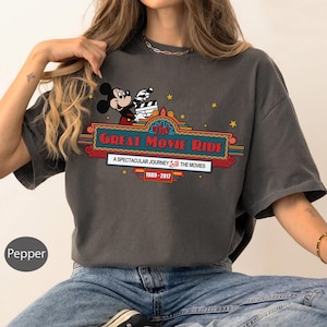 Puede incluir: Camiseta gris oscuro con Mickey Mouse sosteniendo una claqueta de cine. El texto "The Great Movie Ride" se muestra en un dise&ntilde;o de marquesina colorido, con "A Spectacular Journey Into The Movies 1989-2017".