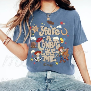 Pu&ograve; includere: Una t-shirt blu con una stampa grafica che riporta il testo "You're A Cowboy Like Me" e "Andy" sulla suola di uno stivale. Il design include illustrazioni di Woody, Jessie, Bullseye, una stella da sceriffo, un ferro di cavallo e un lazo.