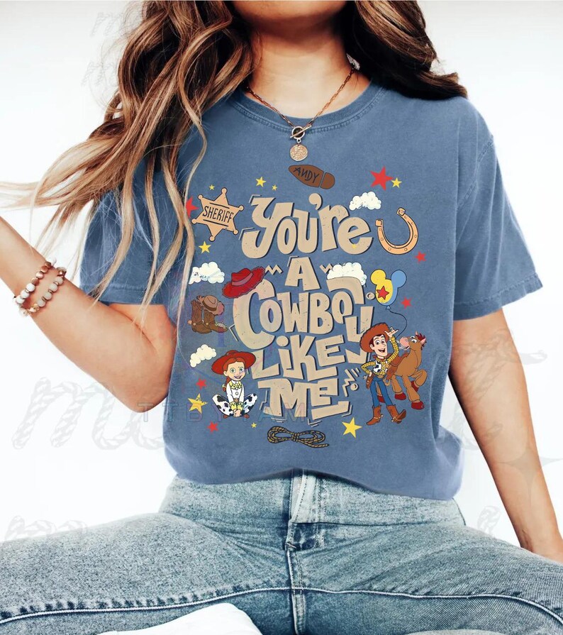 Pu&ograve; includere: Una t-shirt blu con una stampa grafica che riporta il testo "You're A Cowboy Like Me" e "Andy" sulla suola di uno stivale. Il design include illustrazioni di Woody, Jessie, Bullseye, una stella da sceriffo, un ferro di cavallo e un lazo.