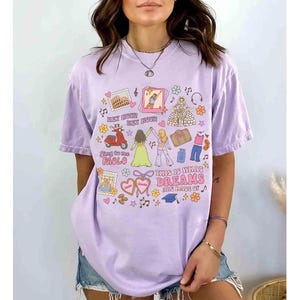 K&ouml;nnte beinhalten: Ein lila T-Shirt mit einem farbenfrohen Grafikdesign. Das Design beinhaltet Illustrationen eines Rollers, Wahrzeichen und den Text "THIS IS WHAT DREAMS ARE MADE OF". Das Shirt hat kurze &Auml;rmel und eine lockere Passform.