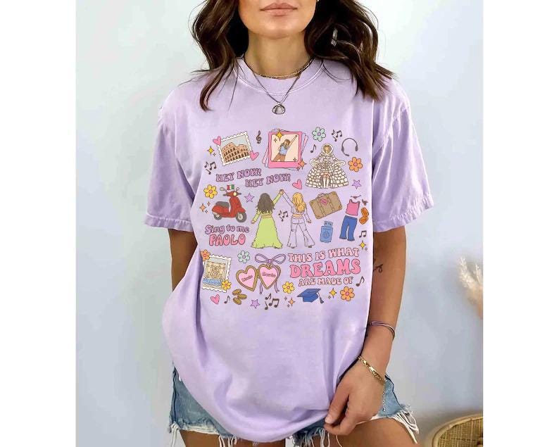 K&ouml;nnte beinhalten: Ein lila T-Shirt mit einem farbenfrohen Grafikdesign. Das Design beinhaltet Illustrationen eines Rollers, Wahrzeichen und den Text "THIS IS WHAT DREAMS ARE MADE OF". Das Shirt hat kurze &Auml;rmel und eine lockere Passform.