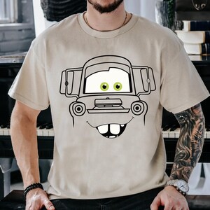May include: A beige short-sleeved t-shirt featuring a large black line-art graphic of a cartoon tow truck face. The design includes prominent white eyes with green pupils, a wide smiling mouth with two visible front teeth, and a truck hood.