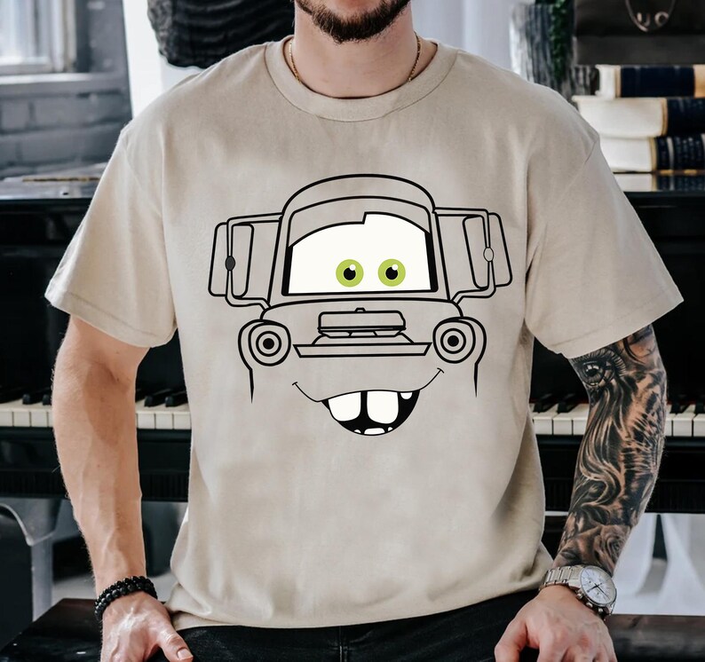 May include: A beige short-sleeved t-shirt featuring a large black line-art graphic of a cartoon tow truck face. The design includes prominent white eyes with green pupils, a wide smiling mouth with two visible front teeth, and a truck hood.