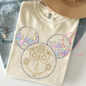 May include: Cream-colored Comfort Colors t-shirt featuring a whimsical design of a smiling face with circular elements. The design includes colorful illustrations of buildings and structures. The shirt is paired with blue jeans and a necklace.