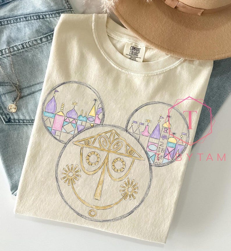 May include: Cream-colored Comfort Colors t-shirt featuring a whimsical design of a smiling face with circular elements. The design includes colorful illustrations of buildings and structures. The shirt is paired with blue jeans and a necklace.