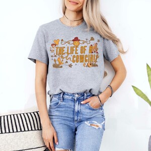 May include: A heather gray t-shirt with the text "THE LIFE OF A COWGIRL" in a western-themed design. The graphic includes a cowboy hat, boots, and stars. The shirt is paired with blue denim shorts.