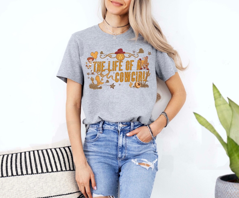 May include: A heather gray t-shirt with the text "THE LIFE OF A COWGIRL" in a western-themed design. The graphic includes a cowboy hat, boots, and stars. The shirt is paired with blue denim shorts.