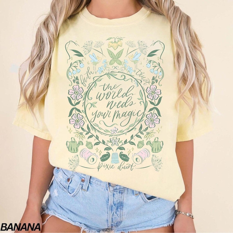 Pu&ograve; includere: T-shirt giallo chiaro con la scritta "the world needs your magic" circondata da illustrazioni floreali e botaniche. La maglietta presenta anche le parole "faith", "trust" e "pixie dust".