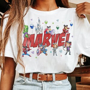 May include: White t-shirt featuring the word "MARVEL" in red block letters, with cartoon illustrations of Marvel characters. The design includes a castle, balloons, and stars. The shirt has a crew neck and short sleeves.