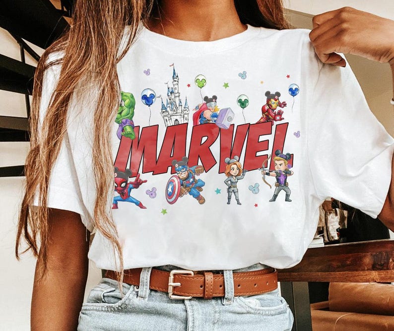 May include: White t-shirt featuring the word "MARVEL" in red block letters, with cartoon illustrations of Marvel characters. The design includes a castle, balloons, and stars. The shirt has a crew neck and short sleeves.
