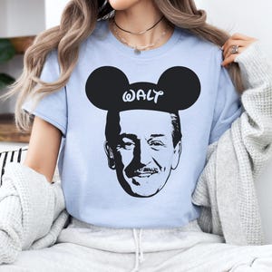 Puede incluir: Camiseta azul claro con un gr&aacute;fico en blanco y negro de Walt Disney con orejas de Mickey Mouse. La palabra "WALT" est&aacute; impresa encima de la imagen. La camiseta es de cuello redondo y manga corta.