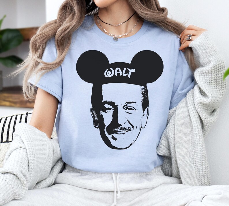 Puede incluir: Camiseta azul claro con un gr&aacute;fico en blanco y negro de Walt Disney con orejas de Mickey Mouse. La palabra "WALT" est&aacute; impresa encima de la imagen. La camiseta es de cuello redondo y manga corta.