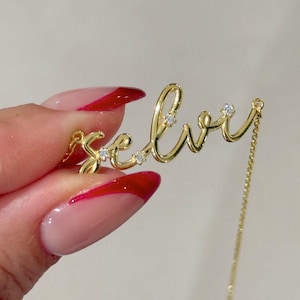 May include: A gold-toned necklace featuring a cursive nameplate pendant that reads Selvi. The script-style lettering is accented with four small, clear, round-cut stones embedded within the metal surface, attached to a delicate gold-toned chain.