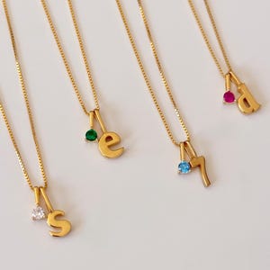 May include: Four gold-toned box chain necklaces, each featuring a small pendant consisting of a lowercase letter or number paired with a single round-cut gemstone. The charms include the letter S with a clear stone, e with a green stone, 7 with a blue stone, and d with a pink stone.