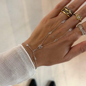 May include: A silver hand chain bracelet with rectangular, clear stones. The bracelet connects to a ring and wraps around the hand. Several rings with gold and clear stones are also visible.