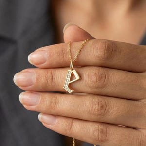 May include: A gold-colored pendant necklace featuring the letter "A" encrusted with small, clear gemstones. The pendant hangs from a delicate gold chain. The necklace is held up by a person's hand.
