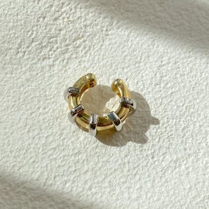 May include: A gold-toned ear cuff with silver accents. The ear cuff has a rounded shape with a thick band and is adorned with multiple silver-colored rings. The design is simple yet elegant, suitable for everyday wear.