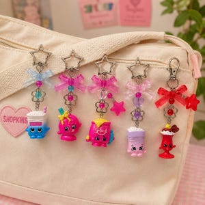 May include: Five colourful keychains featuring Shopkins characters, each attached to a silver star-shaped clasp. The charms include a blue and white drink, a pink circular character, a pink flour bag, a purple container, and a red sundae, all accented with bows.
