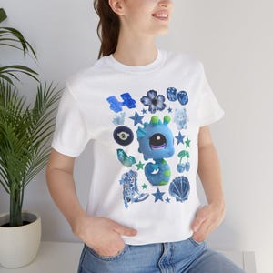 Puede incluir: Camiseta blanca con un diseño de dragón azul caprichoso y varios elementos azules y verdes. El gráfico incluye estrellas, flores, dados y otros elementos lúdicos. La camiseta está hecha de un material suave.