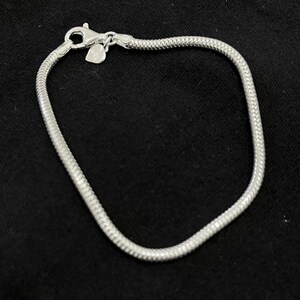 Puede incluir: Una pulsera plateada con un diseño de cadena de serpiente. La pulsera tiene un cierre de mosquetón y un pequeño dije en forma de corazón. La pulsera se muestra sobre una superficie negra.