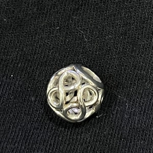 May include: A small, round, silver-colored charm with an openwork design of intertwined loops. The charm is set against a black background, highlighting its intricate details and metallic sheen. This decorative charm could be used for jewelry or other crafts.