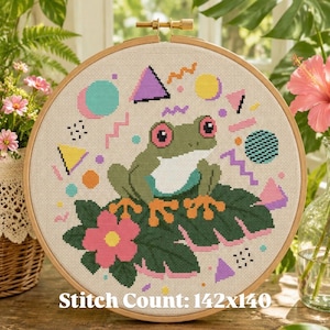 May include: Cross-stitch embroidery of a green frog with pink-rimmed eyes, sitting on a leaf with a pink flower. The design includes geometric shapes in pastel colors. The text "Stitch Count: 142x140" is at the bottom.