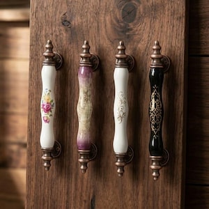 May include: Four decorative cabinet pulls with ornate designs. The pulls feature white, black, and purple ceramic handles with floral and scroll patterns. The hardware is a bronze color, mounted on a wooden surface.